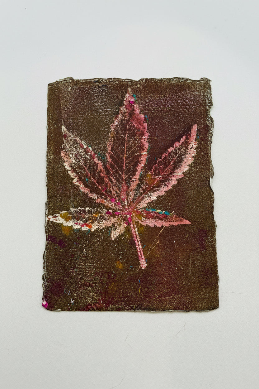 A square textured art print or painting featuring a pink cannabis leaf on a dark brown background, displayed flat against a light off-white surface.