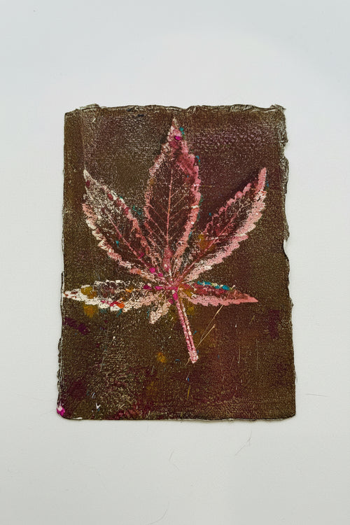 A square textured art print or painting featuring a pink cannabis leaf on a dark brown background, displayed flat against a light off-white surface.