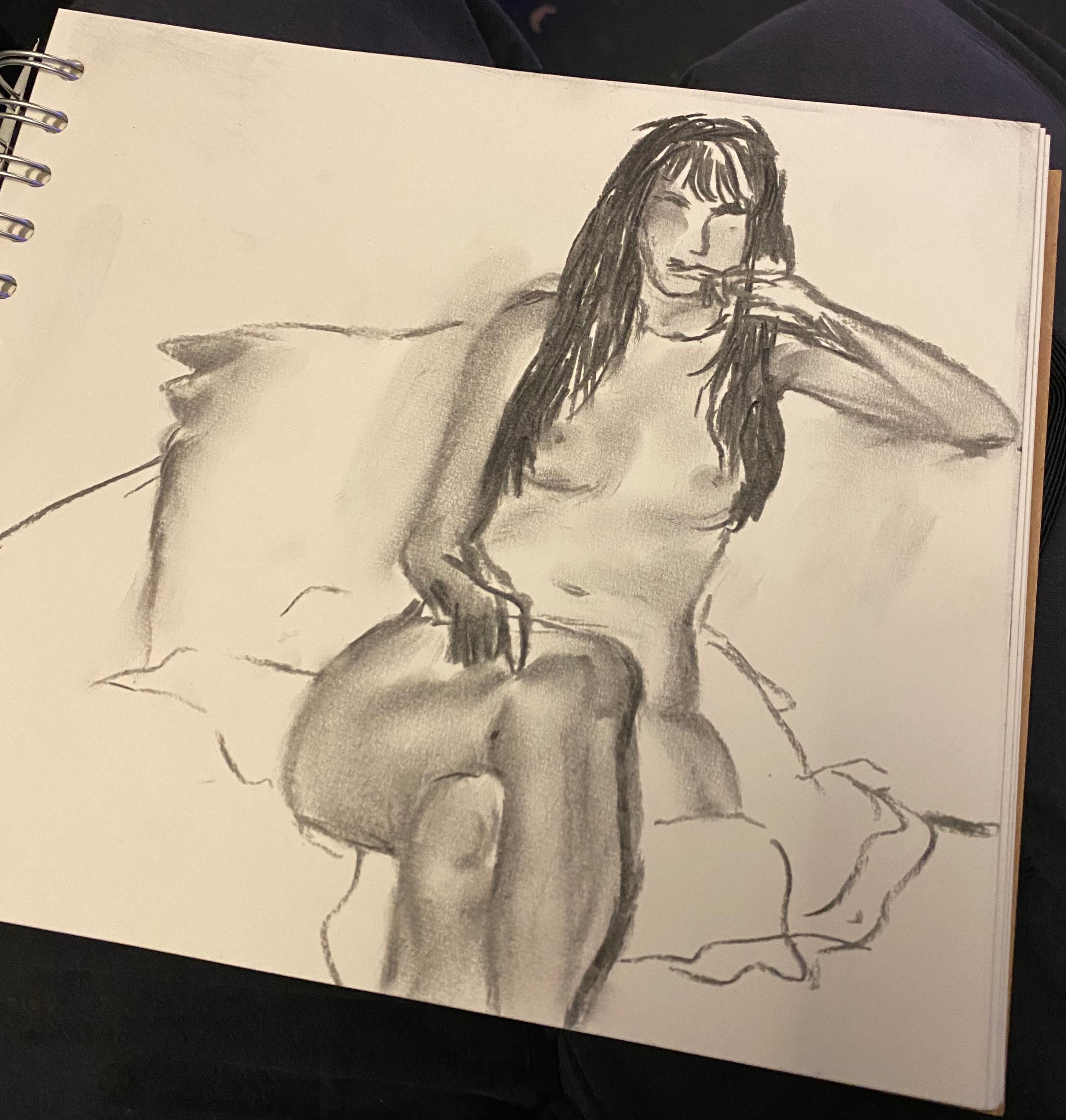 A pencil sketch of a seated nude woman on a spiral-bound drawing page, photographed against a dark background.